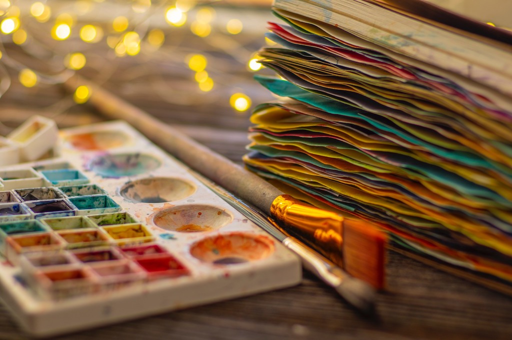 Paints, paintbrushes, and a colorful journal are arranged on a table